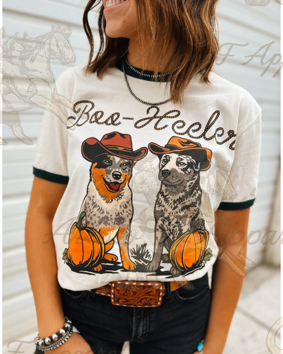 A woman wearing a white graphic t-shirt with 'Baa-Heeler' text and two illustrated dogs wearing cowboy hats with pumpkins. The t-shirt has dark green trim on the sleeves and collar, paired with dark jeans and a brown leather belt.