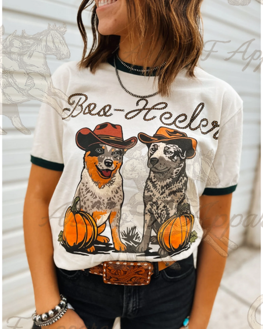 A woman wearing a white graphic t-shirt with 'Baa-Heeler' text and two illustrated dogs wearing cowboy hats with pumpkins. The t-shirt has dark green trim on the sleeves and collar, paired with dark jeans and a brown leather belt.