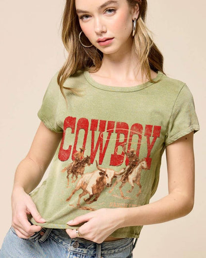 Woman wearing a green t-shirt with 'Cowboy' and horse graphics, paired with denim shorts on a beige background.
